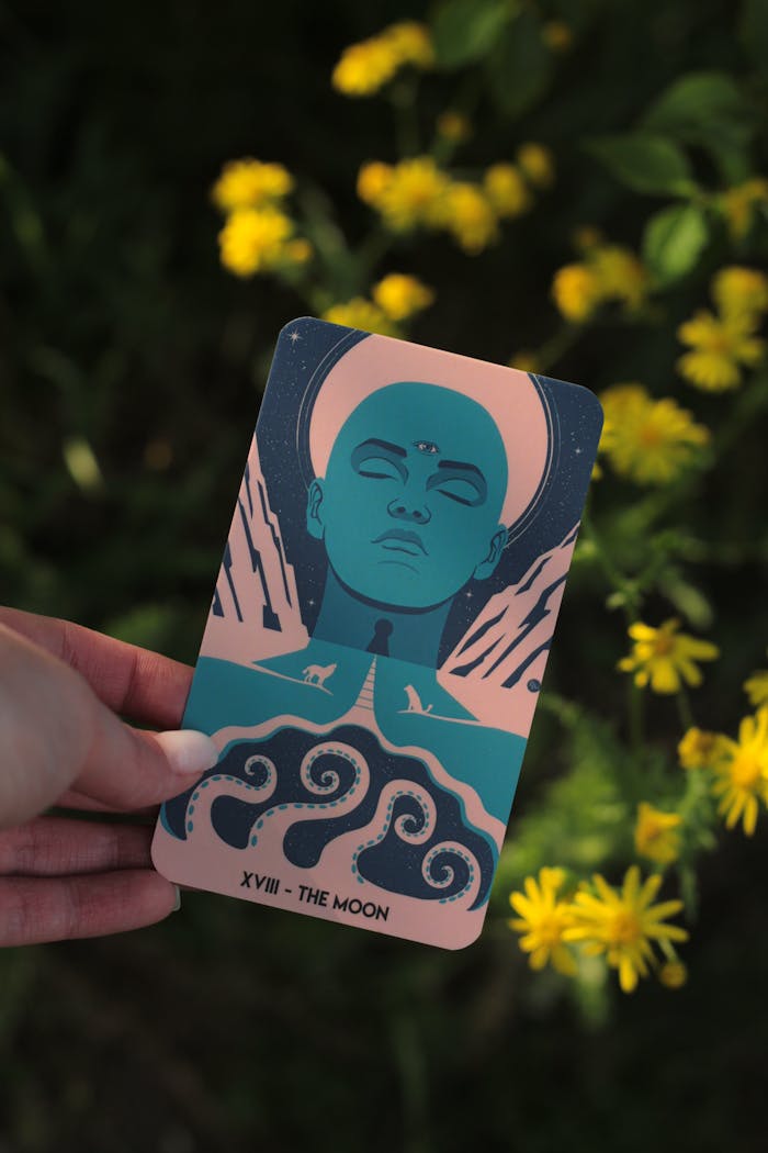 A tarot card featuring 'The Moon' held against a backdrop of yellow flowers in sunlight.